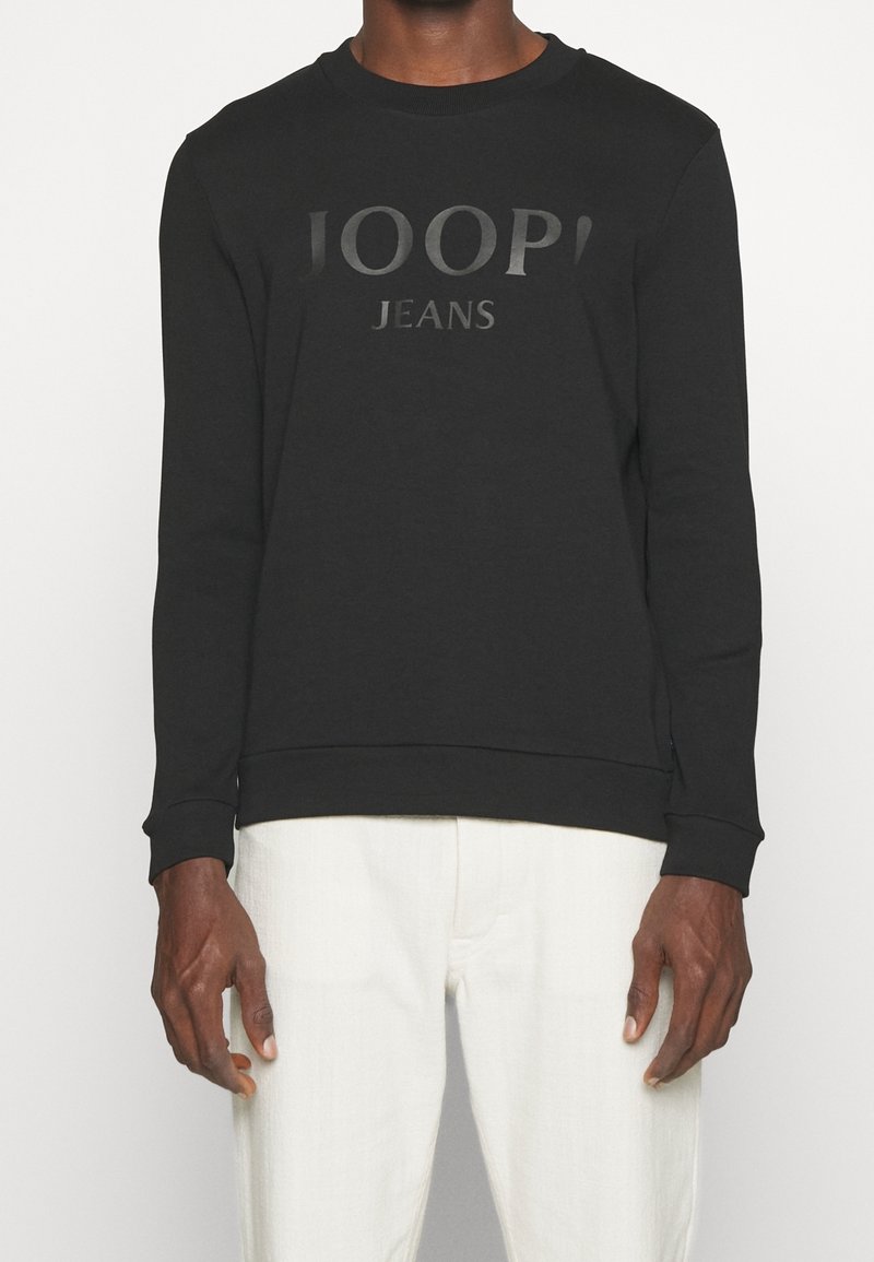 Homme portant un pull noir à manches longues avec le texte "JOOP! JEANS" et un pantalon blanc, debout devant un fond uni.