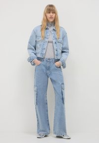 Giacca di jeans blu chiaro e pantaloni cargo a gamba larga, caratterizzati da molteplici tasche e una morbida maglia grigia. Il tessuto ha una texture leggermente usurata.