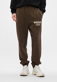 Καφέ φόρμες μ�ε λάστιχο στη μέση, υφασμάτινη υφή και εκτυπωμένο λογότυπο σε μπεζ. Συνδυάζονται με άσπρα αθλητικά παπούτσια.