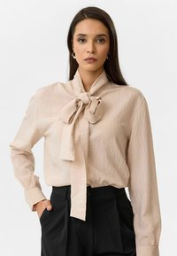 Lichtbeige blouse met een gestructureerde afwerking, voorzien van een grote strik bij de halslijn en lange mouwen, gecombineerd met zwarte op maat gemaakte broeken.