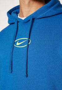 Modrá bavlněná mikina s kapucí a šňůrkami, se žlutým logem Nike na hrudi. Hladká textura a uvolněný střih.