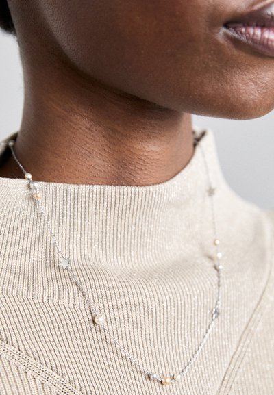 Collar de plata con acentos de estrella y cuentas, exhibido sobre una blusa beige texturizada, que presenta un brillo sutil.