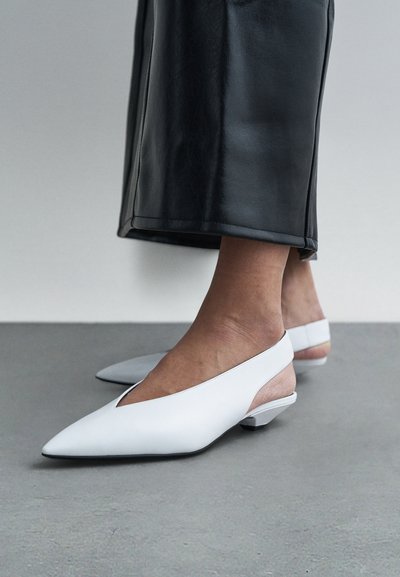 Baltas ādas kurpes ar atvērumu aizmugurē, tapas formu un zemiem papēžiem, ar izgriezuma dizainu un gludu tekstūru uz betona virsmas.