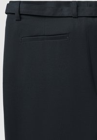 Pantaloni neri su misura realizzati in tessuto liscio, con vita piatta, vestibilità slim e una tasca posteriore singola. Accentuati da una cintura coordinata.