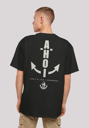 Black t-shirt featuring a large white graphic print with "AHOI," arrows, and the text “Knut & Jan | Hamburg” in a bold font.