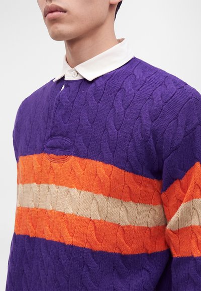 Homme portant un pull violet en maille torsadée avec des rayures horizontales orange et beige par-dessus une chemise blanche à col.