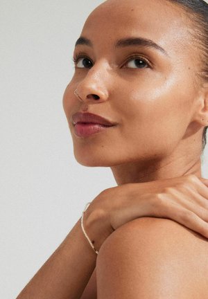 Close-up of a face with smooth, glowing skin, dark hair pulled back, and a silver nose ring. A delicate bracelet accents the wrist.
