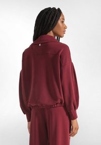 Giacca pullover bordeaux con vestibilità rilassata, maniche lunghe e vita elasticizzata. Tessuto morbido e leggero con una leggera lucentezza.