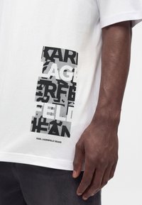 Κοντινή λήψη ενός λευκού t-shirt με μαύρο αφηρημένο κειμενικό σχέδιο και "KARL LAGERFELD JEANS" τυπωμένο κοντά στο στρίφωμα, που φοριέται από άτομο με σκούρο δέρμα.