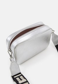 Sömlös silverfärgad lädercrossbodyväska med texturerad yta, två zippered-fack och ett svart justerbart band med logomönster.