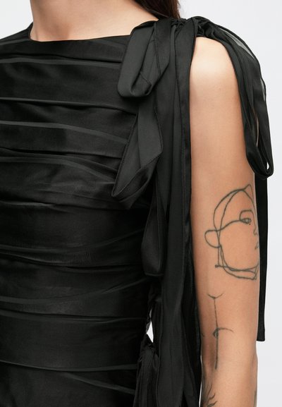 Čierne šaty s vertikálnymi záhybmi a ramennými viazačkami pripomínajúcimi stuhu. Hladký materiál s detailným tetovaním na odkrytej ruke.