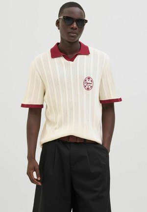 Knit polo shirt in cream with a maroon collar and trim. Features a decorative embroidered patch and a loose fit for comfort.