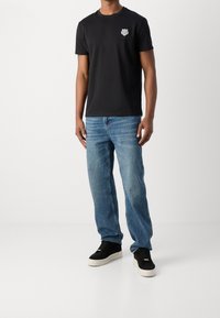 Black cotton t-shirt with round neckline and small logo; paired with light blue denim jeans and black sneakers with white soles.