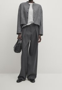 Grijze cropped jacket met zwarte knopen, over een lichtgrijs shirt. Donkergrijze wijde broek en een geweven zwarte handtas. Zwarte schoenen.