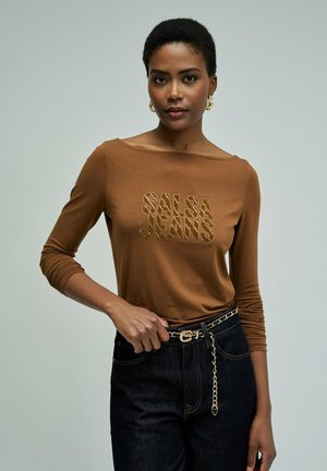 Mujer con el cabello corto que lleva una camisa de manga larga marrón con el texto "SALSA JEANS" y jeans oscuros con un cinturón de cadena dorada sobre un fondo gris.