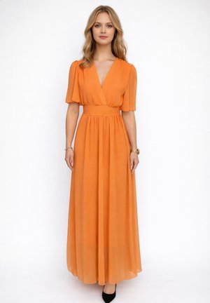 Femme portant une robe longue orange à manches courtes et col en V, debout devant un fond blanc uni.