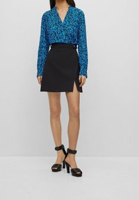 Kvinna som bär en blåmönstrad långärmad blus instoppad i en svart minikjol med sidoval och svarta högklackade sandaler.