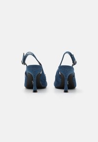 Blå denim slingbackklackar med en böjd silhuett, slanka svarta klackar och justerbar ankelrem som visar en texturerad tygfinish.