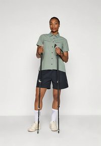 Abbigliamento da escursionismo composto da una camicia verde chiaro a maniche corte con bottoni e pantaloncini scuri, abbinati a bastoncini da trekking e scarpe beige.