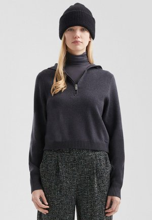 Junge Frau trägt schwarze Strickmütze, dunklen Reißverschluss-Pullover über Rollkragenpullover und strukturierte, hoch taillierte graue Hose, steht vor schlichtem Hintergrund.