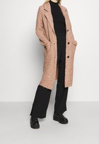 Abrigo largo texturizado en tonos rosa y naranja, con un ajuste relajado, botones grandes y Bolsillos laterales. Lleva puesto un suéter de cuello alto negro y pantalones negros.