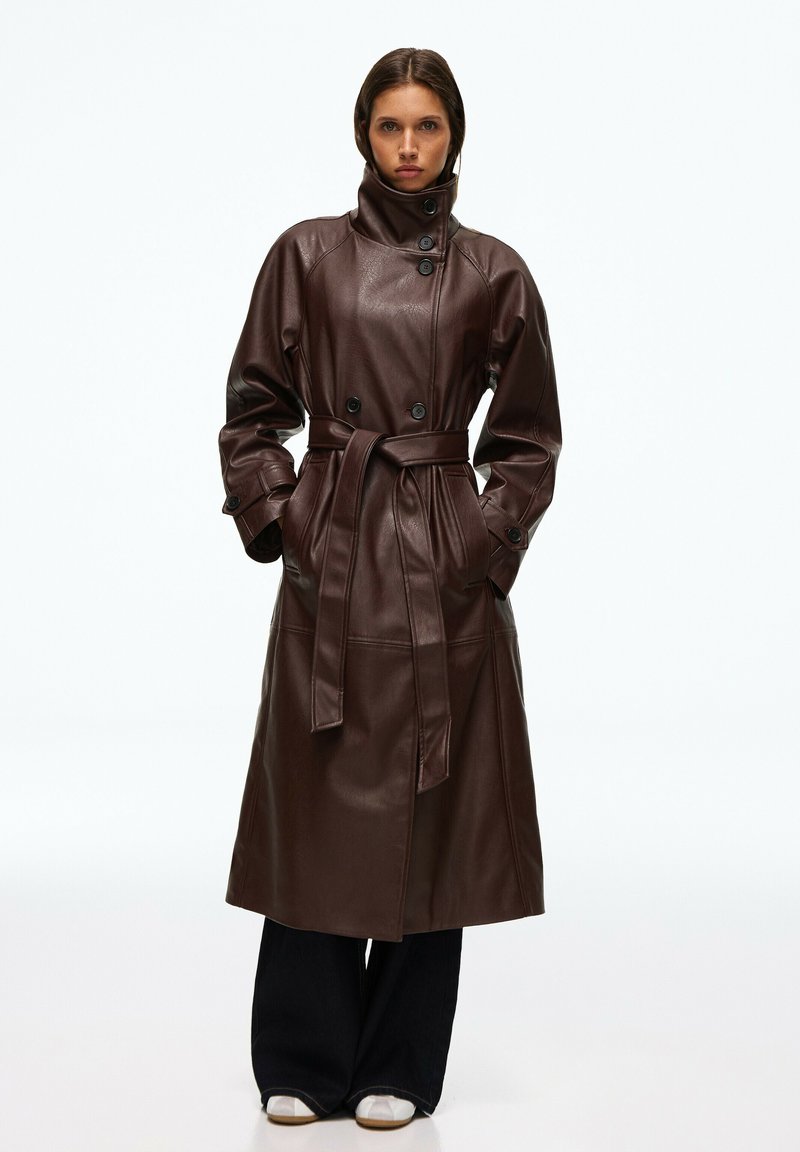 Femme portant un long trench-coat en cuir marron foncé avec col haut et ceinture, pantalon noir à jambes larges et chaussures blanches, debout les mains dans les poches.