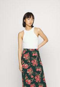 Top branco sem mangas com um decote alto, combinado com uma saia longa verde apresentando padrões florais vermelhos vibrantes e uma textura suave.