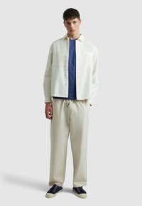 Camicia bianca con bottoni sopra una maglietta blu, abbinata a pantaloni crema e sneakers blu scuro. La camicia ha due tasche sul petto e un taglio rilassato.