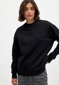 Sudadera negra con un cuello alto, mangas largas y puños acanalados. Tejido suave con un corte relajado y sin patrones ni acentos visibles.