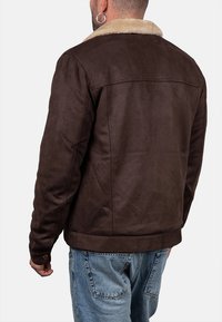 Uomo che indossa una giacca in suede marrone con un colletto in shearling beige e jeans in denim blu, visto da dietro su uno sfondo bianco.