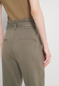 Olive green trousers with a smooth texture, featuring a wide waistband, two back pockets, and a relaxed fit. The top is a tan ribbed tank.