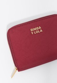 Bimba y Lola cartera burdeos en tejido texturizado, con un cierre dorado y detalles de logo. Forma rectangular compacta.