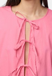 Top de algodón rosa con cuello redondo y lazos, diseño abierto en el frente, que muestra una textura suave y acentos minimalistas.
