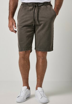 Mand iført olivengrønne shorts med snøre og sidelommer samt hvide lave sneakers, stående mod en ensfarvet baggrund.