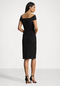 Melns off-shoulder kleita ar pieguļošu siluetu, līdz ceļgaliem, ar rāvējslēdzēju aizmugurē; valkāta ar melniem siksniņu augstiem papēžiem.