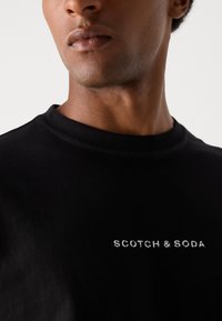 Black crewneck sweatshirt made of smooth cotton, featuring white embroidered text "SCOTCH & SODA" on the chest. Minimalist design.