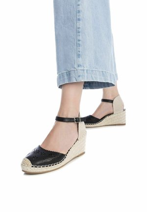 Zapatos de cuña de cuero negro con diseño floral recortado, tacón de yute tejido y fina correa ajustable en el tobillo. Combinados con pantalones de mezclilla azul claro.