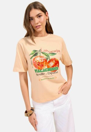 Femme portant un t-shirt beige avec deux motifs de boules disco orange et le texte "Las Naranjas, Vacaciones, Seville España", assorti à un pantalon blanc.