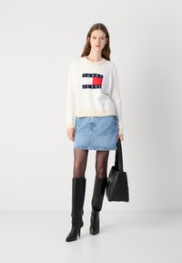 Kremgenser med marineblå og rød logo, lyseblå denimskjørt, svarte knelang støvletter og en svart veske. Glatte teksturer, avslappet stil.