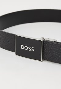 Schwarzer Leder-Gürtel mit strukturiertem Finish, ausgestattet mit einer rechteckigen silbernen Schnalle, die das Wort "BOSS" eingraviert hat.