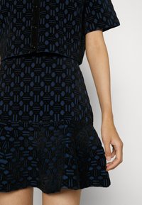 Gros plan d'une personne portant un haut boutonné à motifs noir et bleu foncé et une jupe évasée assortie, avec un bras nu visible.