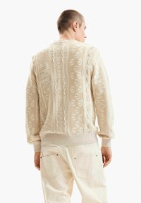 Rövid hajú férfi, aki krémszínű, texturált kötött pulóvert és világos bézs nadrágot visel, hátrafelé néz, fehér háttér előtt.