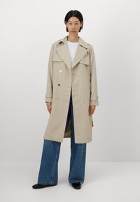 Beige trench coat with double-breasted gold buttons, large lapels, and shoulder epaulets. Worn over a white shirt and paired with wide-leg jeans.