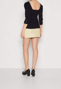 Schwarzes, geripptes Oberteil mit quadratischem Ausschnitt, Dreiviertelärmeln und Knopfakzenten, kombiniert mit einem hellbeigen, strukturierten Minirock und hochhackigen Schuhen.