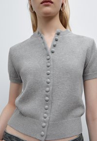 Top grigio a maniche corte in maglia con scollatura a V, caratterizzato da bottoni frontali e orlo a coste. Il materiale sembra morbido con una texture liscia.