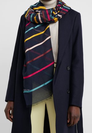 Manteau foncé sur un col roulé clair et un pantalon beige, avec une écharpe rayée colorée enroulée autour du cou et tombant sur le devant.