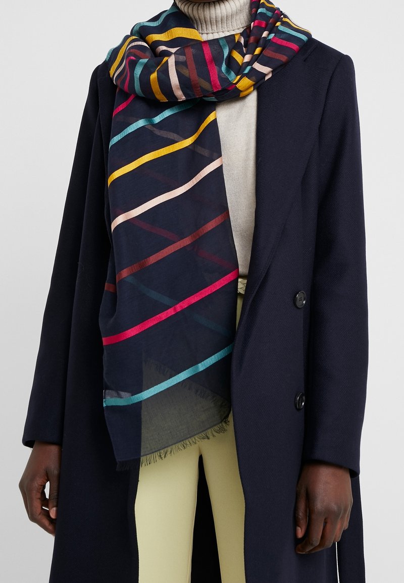 Manteau foncé sur un col roulé clair et un pantalon beige, avec une écharpe rayée colorée enroulée autour du cou et tombant sur le devant.