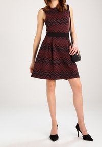 Une robe sans manches, arrivant au genou, avec un motif en zigzag rouge et noir, présentant un corsage ajusté et une jupe évasée. Portée avec des talons noirs et un sac bandoulière.