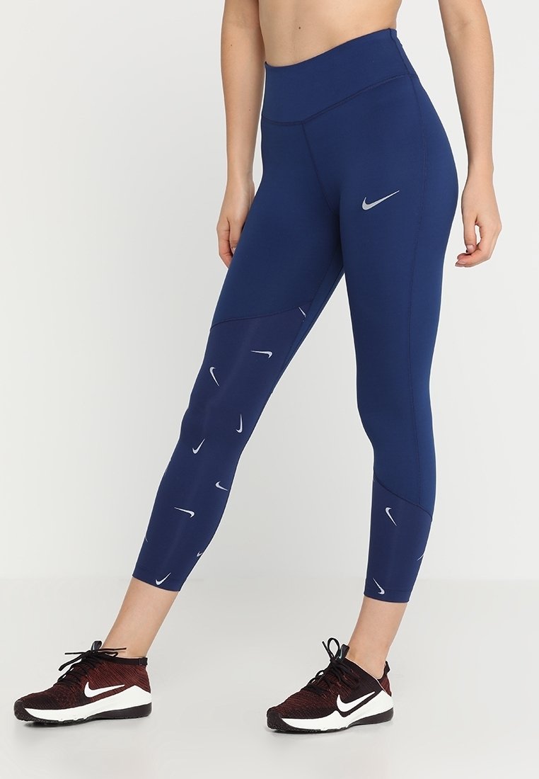 Personne portant un legging Nike bleu marine taille haute avec plusieurs petits logos sur une jambe et des baskets Nike bordeaux avec semelles blanches.
