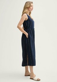 Marineblaues Midi-Kleid mit weißen Punkten, ärmellos, mit tailliertem Bund, seitlichen Taschen und Schlitzen; aus leichtem, weichem Stoff gefertigt.